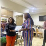 At a doctor's office in Abuja, a woman proudly holds a certificate in front of a group of people in doctor office in Abuja